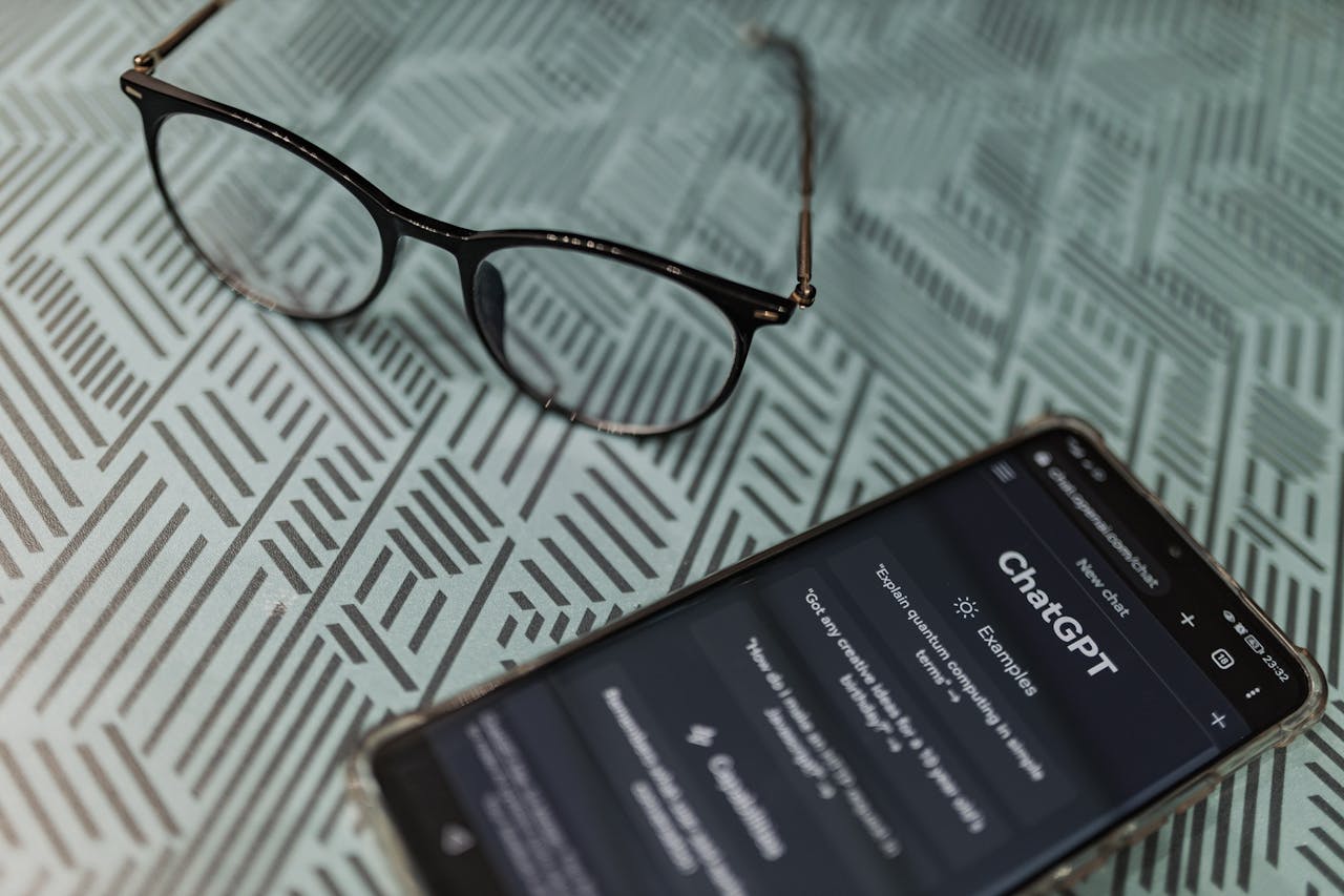 gallery-2 Eyeglasses next to a smartphone displaying the ChatGPT AI app on a patterned surface.